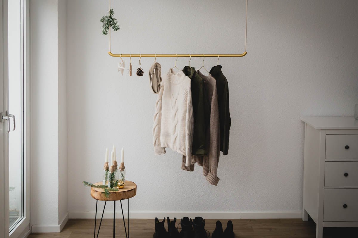 In einem minimalistischen Raum hängen Pullover in neutralen Farben elegant auf Kleiderbügeln aus stabilem Metall von rod & knot – 8 Stück entlang eines eleganten goldenen Gestells. Darunter verleiht ein Holztisch mit brennenden Kerzen und Kieferndekor dem Ambiente Wärme. Schwarze Schuhe sind ordentlich angeordnet, während natürliches Licht durch das Fenster hereinströmt und die ruhige Atmosphäre verstärkt.